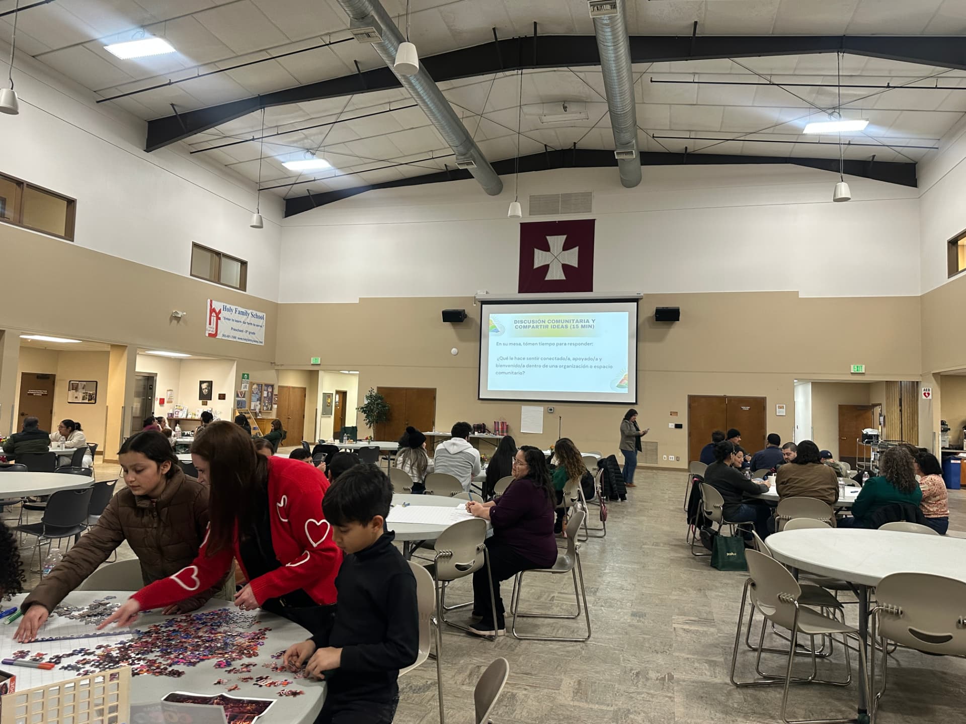 Encuentro comunitario con familias en varias mesas y una pantalla de presentación al centro